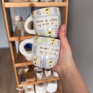 Könnte beinhalten: Zwei Keramikbecher mit Henkeln, mit handgemaltem Design. Die Becher haben eine cremefarbene Basis mit einem sich wiederholenden Muster aus Häusern, Schlüsseln und Blumen. Der Text "home is where my mom is" steht auf der Vorderseite jedes Bechers.