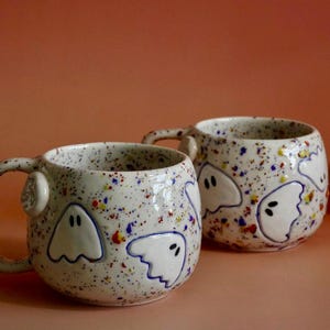 May include: Two round ceramic mugs with a speckled white glaze featuring raised white ghost figures with blue outlines. The mugs have a rounded handle and a colorful paint splatter pattern in red, blue, and yellow across the exterior surface.