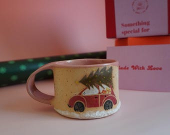 May include: A ceramic mug with a whimsical design featuring a red car carrying a Christmas tree. The mug has a light yellow base with a pink handle and rim. The words "Made With Love" are visible on a card.