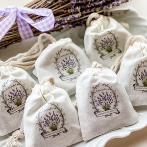 May include: Six small white fabric pouches with a lavender design and the text "Lavender Herbes de Provence" on a white plate. The pouches are tied with a string and are filled with lavender.