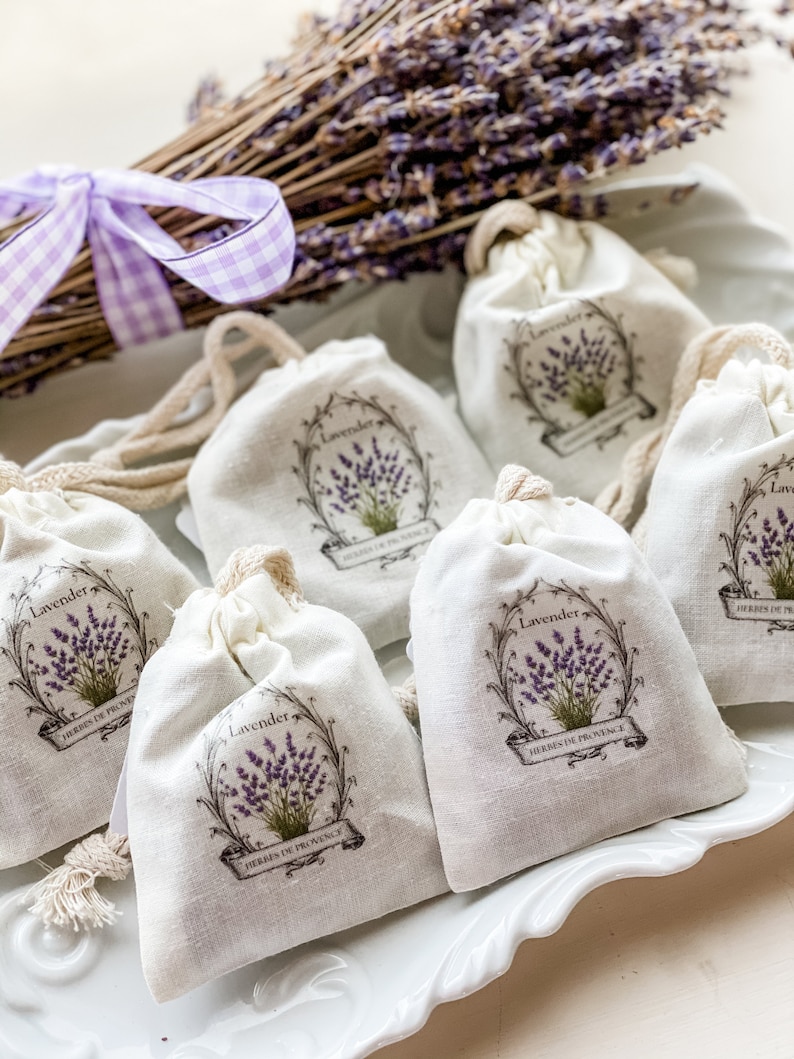 May include: Six small white fabric pouches with a lavender design and the text "Lavender Herbes de Provence" on a white plate. The pouches are tied with a string and are filled with lavender.