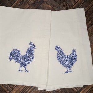 May include: Two white kitchen towels with blue embroidered designs of a rooster and a hen. The designs are made up of floral patterns.