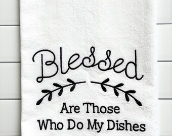 May include: White kitchen towel with the embroidered text "Blessed Are Those Who Do My Dishes" in black script and a decorative leaf design. The towel is hanging against a white tile background.