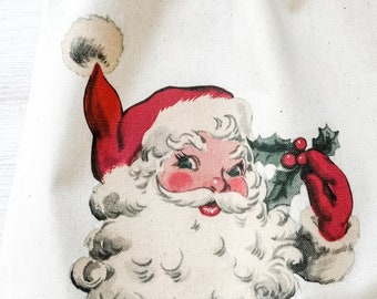 May include: A cream-colored drawstring bag featuring a vintage illustration of Santa Claus. Santa is depicted with a red hat and coat, a white beard, and rosy cheeks. He is holding holly with red berries. The bag is made of a textured fabric.