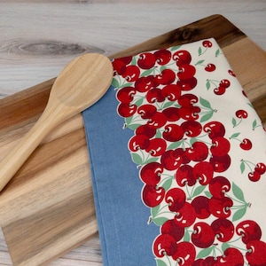 May include: A wooden spoon rests on a wooden cutting board next to a blue and white kitchen towel. The towel features a repeating pattern of bright red cherries with green leaves. The towel is folded, showing both the cherry pattern and the solid blue fabric.