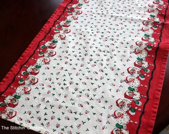 Op de afbeelding: Een wit tafelkleed met een rode rand en een herhalend patroon van groene hulstbladeren en kerstmangezichten. De tekst "The Stitch'n Chicken" is gedrukt in de linkerbenedenhoek.