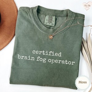 Może przedstawiać: Koszulka Comfort Colors w kolorze mchu z białym napisem "certified brain fog operator". Złoty naszyjnik z wisiorkiem jest ułożony na koszulce. Koszulka leży na białej powierzchni.