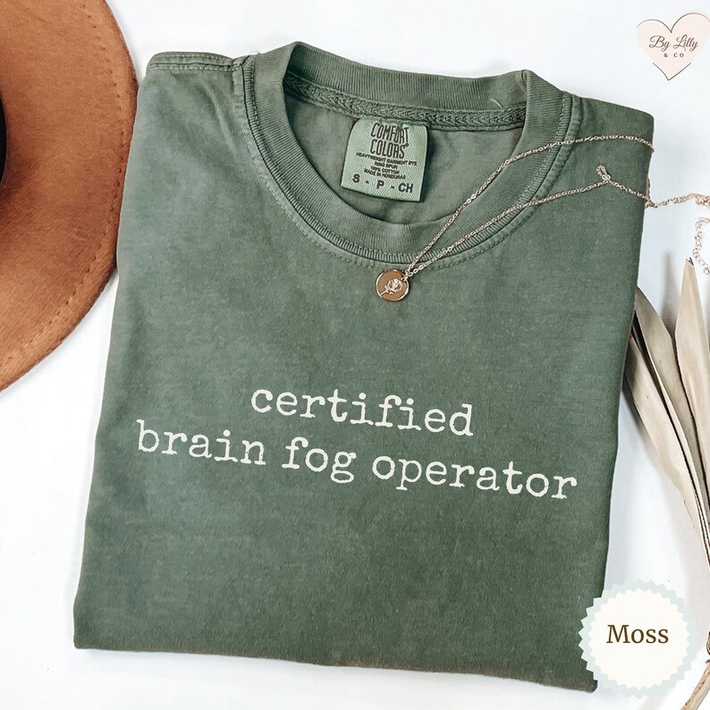 Może przedstawiać: Koszulka Comfort Colors w kolorze mchu z białym napisem "certified brain fog operator". Złoty naszyjnik z wisiorkiem jest ułożony na koszulce. Koszulka leży na białej powierzchni.