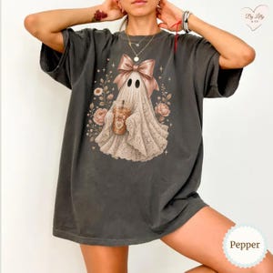 May include: A charcoal gray short-sleeve t-shirt featuring a graphic of a ghost wearing a large tan bow and holding a coffee cup. The ghost is surrounded by tan floral illustrations and small star shapes. The word Pepper is printed in the corner.