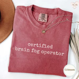 Może przedstawiać: Koszulka Comfort Colors w kolorze karmazynowym z białym napisem "certified brain fog operator". Srebrny naszyjnik z wisiorkiem jest zarzucony na koszulkę. Koszulka jest złożona i ma metkę.