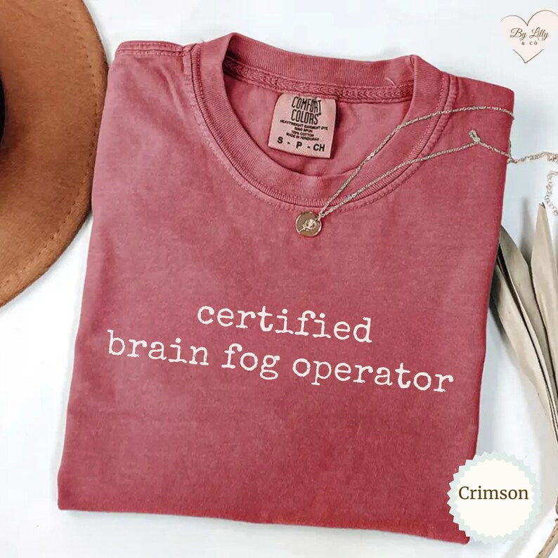 Może przedstawiać: Koszulka Comfort Colors w kolorze karmazynowym z białym napisem "certified brain fog operator". Srebrny naszyjnik z wisiorkiem jest zarzucony na koszulkę. Koszulka jest złożona i ma metkę.