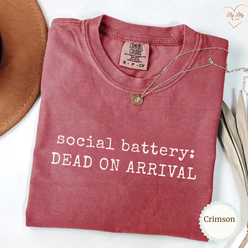 Puede incluir: Camiseta color carmes&iacute; con el texto blanco "social battery: DEAD ON ARRIVAL". La camiseta tiene cuello redondo y una peque&ntilde;a etiqueta. Un collar plateado con un colgante est&aacute; sobre la camiseta.