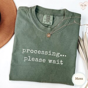 May include: A moss green short-sleeve t-shirt with a crew neckline and the white text "processing... please wait" printed on the front. The shirt features a Comfort Colors brand label inside the collar and a thin gold-toned chain necklace.