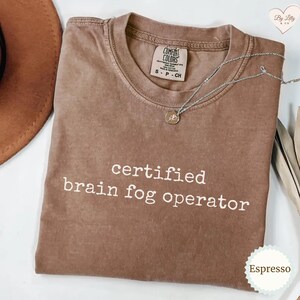 Może przedstawiać: Koszulka w kolorze espresso z białym napisem "certified brain fog operator". Koszulka ma okrągły dekolt i kr&oacute;tkie rękawy. Srebrny naszyjnik z małą zawieszką spoczywa na koszulce. Koszulka jest oznaczona jako "Comfort Colors".