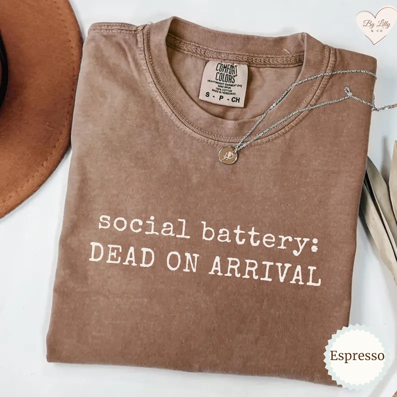 Puede incluir: Camiseta color espresso con el texto blanco "social battery: DEAD ON ARRIVAL". La camiseta tiene cuello redondo y una peque&ntilde;a etiqueta en el cuello. Un collar plateado con un colgante descansa sobre la camiseta.