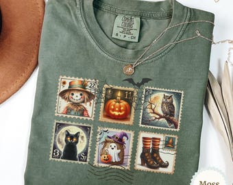 May include: A moss green crewneck t-shirt featuring a graphic print of six vintage-style postage stamps with Halloween imagery, including a scarecrow, a jack-o'-lantern, an owl, a black cat, a ghost, and witch boots, with a spiderweb overlay.