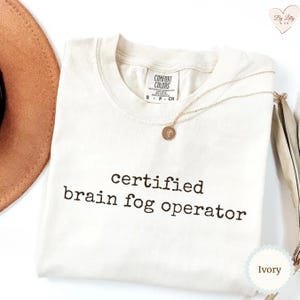 Może przedstawiać: Kremowa koszulka z czarnym napisem "certified brain fog operator". Koszulka ma okrągły dekolt i kr&oacute;tkie rękawy. Złoty naszyjnik z wisiorkiem spoczywa na koszulce. Częściowo widoczny jest brązowy kapelusz.