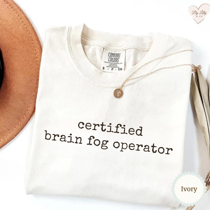 Może przedstawiać: Kremowa koszulka z czarnym napisem "certified brain fog operator". Koszulka ma okrągły dekolt i kr&oacute;tkie rękawy. Złoty naszyjnik z wisiorkiem spoczywa na koszulce. Częściowo widoczny jest brązowy kapelusz.