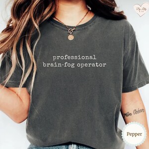 May include: A dark grey t-shirt with the words "professional brain-fog operator" printed in white. The t-shirt has a crew neck and looks to be made of a soft material. A gold necklace is visible at the neckline.