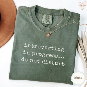 May include: A moss green t-shirt with the text "introverting in progress... do not disturb" in white. The shirt has a crew neck and short sleeves. A gold necklace with a pendant is draped on the shirt.