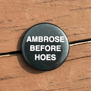 以下が含まれることがあります： 「AMBROSE BEFORE HOES」の白い文字が中央に配置された黒い丸いボタン。ボタンは茶色の木製の表面に置かれています。