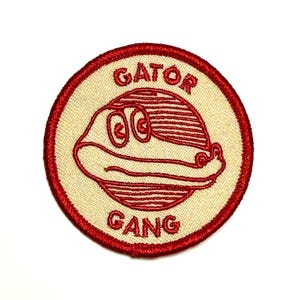 May include: Round embroidered patch with a red border and a cream-colored center. The patch features a red cartoon alligator with the words "GATOR GANG" in red lettering.