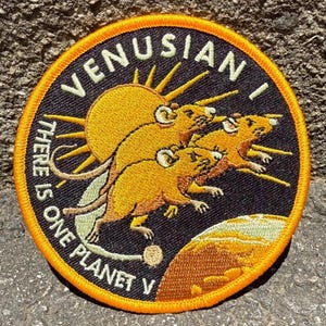 May include: Circular embroidered patch with a black background and an orange border. The patch features the text "VENUSIAN I" and "THERE IS ONE PLANET V". It depicts three yellow rodents in front of a yellow sun.