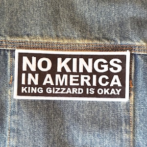 May include: Close-up of a denim jacket with a rectangular patch. The patch is black with white text that reads "NO KINGS IN AMERICA" and "KING GIZZARD IS OKAY". The jacket has visible stitching.