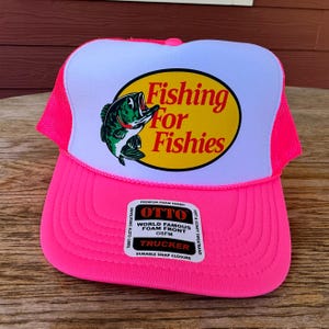 May include: A vibrant pink trucker hat with a white front panel displaying a fish graphic and the words "Fishing For Fishies." The hat has a mesh back and a curved peak. The hat also has a tag that says "OTTO" and "TRUCKER".