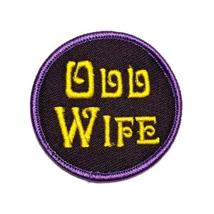 Könnte beinhalten: Ein runder, gestickter Aufnäher mit den Worten "ODD WIFE" in gelben, stilisierten Buchstaben. Der Text ist auf einem schwarzen Hintergrund zentriert, umrahmt von einem lila genähten Rand. Der Aufnäher ist vor weißem Hintergrund isoliert.