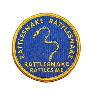 Könnte beinhalten: Ein runder Aufnäher mit blauem Hintergrund und gelbem Rand. Der Aufnäher zeigt ein gelbes Klapperschlangen-Design und den Text "RATTLESNAKE RATTLES ME" in Gelb. Die Worte "RATTLESNAKE" sind oben und unten gebogen.