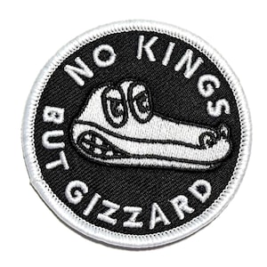 May include: A circular embroidered patch with a black background and a white border. The patch features a white cartoon alligator head in the center and the text NO KINGS BUT GIZZARD embroidered in white capital letters around the perimeter.