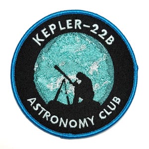 Op de afbeelding: Ronde patch met een zwarte rand en een blauwe buitenrand. De patch heeft de tekst "KEPLER-22B ASTRONOMY CLUB" in het wit. Een silhouet van een persoon die een telescoop gebruikt staat in het midden, met een lichtblauwe achtergrond.