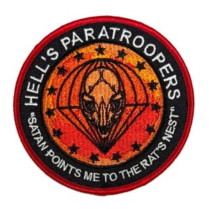 Peut inclure: Écusson brodé circulaire avec une bordure rouge et un dos noir. Le texte "HELL'S PARATROOPERS" et "SATAN POINTS ME TO THE RAT'S NEST" est présent. Le motif central comprend un crâne et un parachute orange et noir.