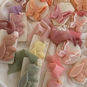 Puede incluir: Una colección de objetos decorativos en colores pastel, que incluyen adornos con forma de mariposa y conejito. Los adornos son en tonos rosa, amarillo, verde y melocotón, con detalles de cinta. Los artículos están dispuestos sobre una superficie blanca texturizada.