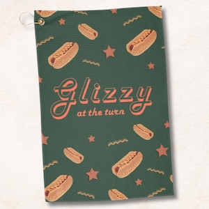 May include: Green golf towel with a pattern of hot dogs and stars. The text "Glizzy at the turn" is printed in orange.