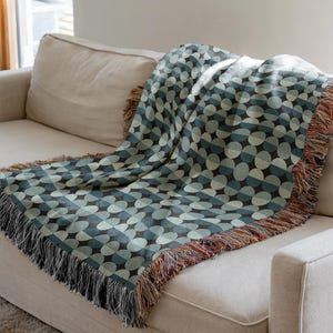 May include: A woven throw blanket featuring a repeating geometric pattern of overlapping circles in shades of dark teal, light blue, and cream. The blanket has a textured finish and is finished with a multi-coloured fringe along the edges.