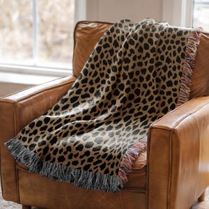 May include: A woven throw blanket featuring a tan background with a repeating black leopard print pattern. The blanket has a textured surface and is finished with decorative fringe along the edges, displaying a mix of black and multi-colored threads.