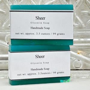 Op de afbeelding: Twee handgemaakte glycerinezeepjes. Elk zeepje is turquoise en heeft een wit etiket met het woord "Sheer" en de tekst "Glycerin Soap Handmade Soap net wt. approx. 99 gram".