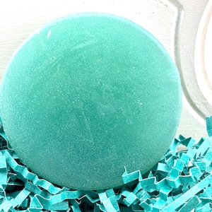 May include: A round, teal-colored soap bar with a textured surface. The soap is sitting on a bed of teal-colored shredded paper.