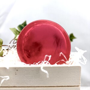 Puede incluir: Un objeto redondo y brillante, de color rojo, posiblemente un jabón o una bomba de baño, descansa en una caja de madera blanca llena de papel triturado blanco. Hojas verdes asoman por detrás del objeto.