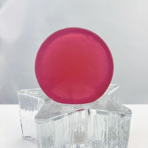 May include: A vibrant, round, magenta-colored object sits atop a clear, star-shaped glass holder. The glass holder has a textured, frosted appearance. The magenta object appears to be solid and opaque.