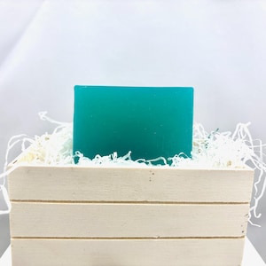 May include: A teal-colored soap bar rests in a small, light-colored wooden crate filled with white paper filler. The soap is rectangular and has a smooth surface. The crate has horizontal slats and is set against a white background.