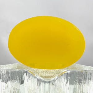 May include: A vivid yellow, oval soap bar sits on a clear, textured glass soap dish. The soap has a smooth surface and a bright colour. The dish has a star-like shape with a rough, icy texture.
