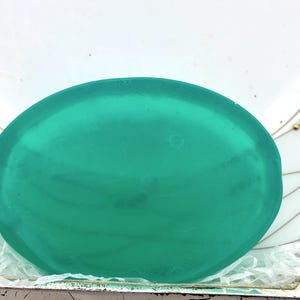 Puede incluir: Un objeto grande, de forma ovalada, de color verde azulado. El objeto tiene una superficie lisa y brillante y parece estar hecho de un material translúcido. El objeto está sobre un fondo blanco con un borde decorativo.