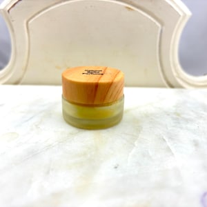 May include: A small glass jar with a wooden lid. The lid has the text "Natural Solid Perfume" printed on it. The jar is filled with a yellow substance.