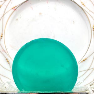 May include: A teal-coloured, semi-spherical object is the focal point. It's set against a white plate with gold and pink decorative lines and gold bead accents. The plate's design adds a touch of elegance.