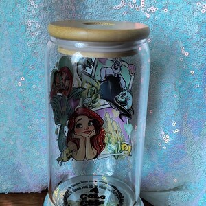 Op de afbeelding: Een heldere glazen pot met een houten deksel, met een kleurrijke afbeelding van Ariel en andere personages uit De Kleine Zeemeermin. De pot staat op een zilveren voet en de achtergrond is een glinsterende, lichtblauwe stof.