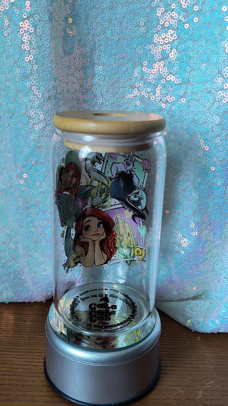 Op de afbeelding: Een heldere glazen pot met een houten deksel, met een kleurrijke afbeelding van Ariel en andere personages uit De Kleine Zeemeermin. De pot staat op een zilveren voet en de achtergrond is een glinsterende, lichtblauwe stof.