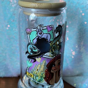 Op de afbeelding: Een doorzichtige glazen beker met een bamboe deksel, met kleurrijke illustraties van personages uit een populaire animatiefilm. De beker staat op een zilveren voet en is ongeveer 15 cm hoog.